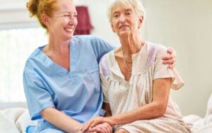 Betreuungskraft sitzt mit demenzkrankem Senior am Küchentisch und spricht ruhig in vertrauter Umgebung