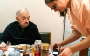 Betreuungskraft serviert einem Senior zuhause eine ausgewogene Mahlzeit am Esstisch