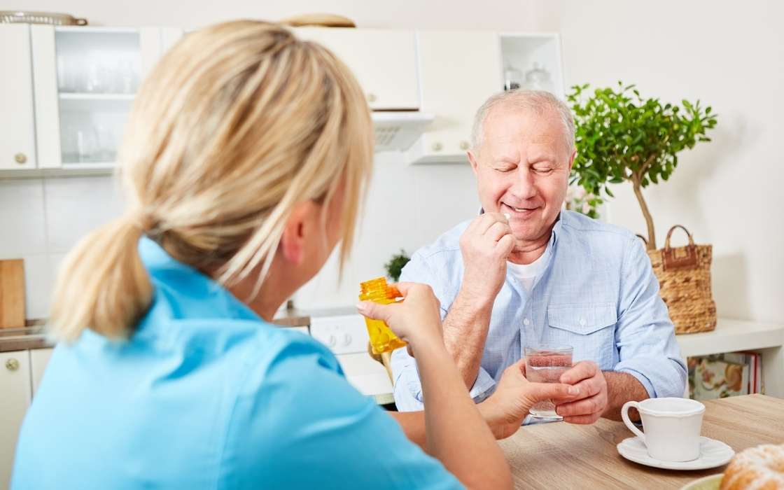 Eine blonde Betreuungskraft in blauer Arbeitskleidung reicht einem lächelnden älteren Herrn am Küchentisch eine gelbe Medikamentenpackung und ein Glas Wasser.